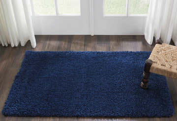 Living room scene with a Nourison Malibu Shag Navy rug on wood floor, white curtains, and a woven stool.