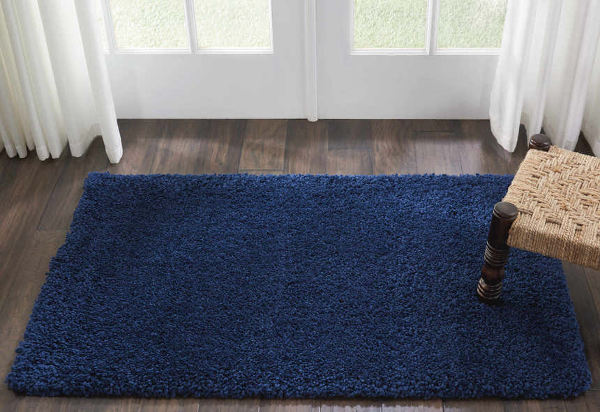 Living room scene with a Nourison Malibu Shag Navy rug on wood floor, white curtains, and a woven stool.