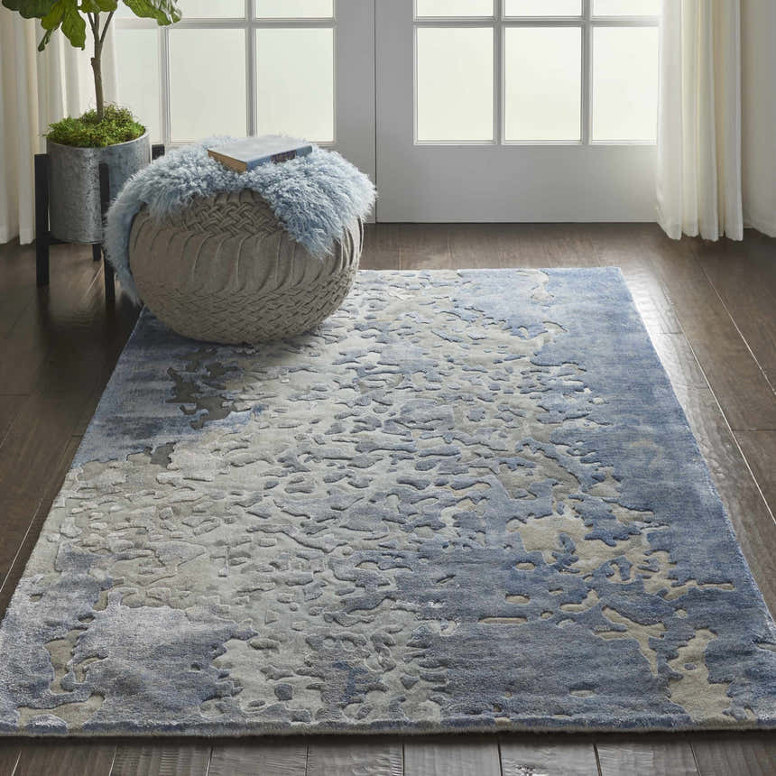 Living room scene with a blue Nourison Prismatic Denim area rug, woven ottoman, wood floors, and natural light.