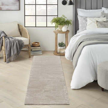 Bedroom scene with a Michael Amini Ma30 Star Taupe/Ivory rug, rattan chair, wood side table, and bed with white linens.