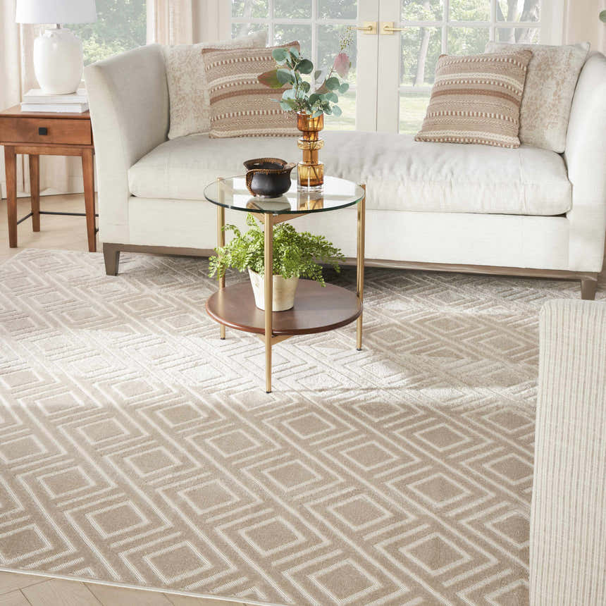 Living room scene with a beige Nourison Care Free rug featuring a geometric diamond pattern. Sofa, glass table, and natural light.