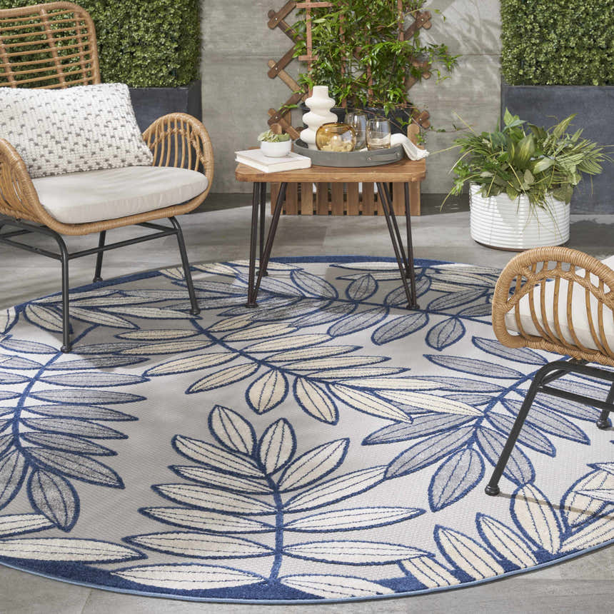 Outdoor patio scene with a round Nourison Aloha Ivory Navy Tropical Area Rug, wicker chairs, and a small table.