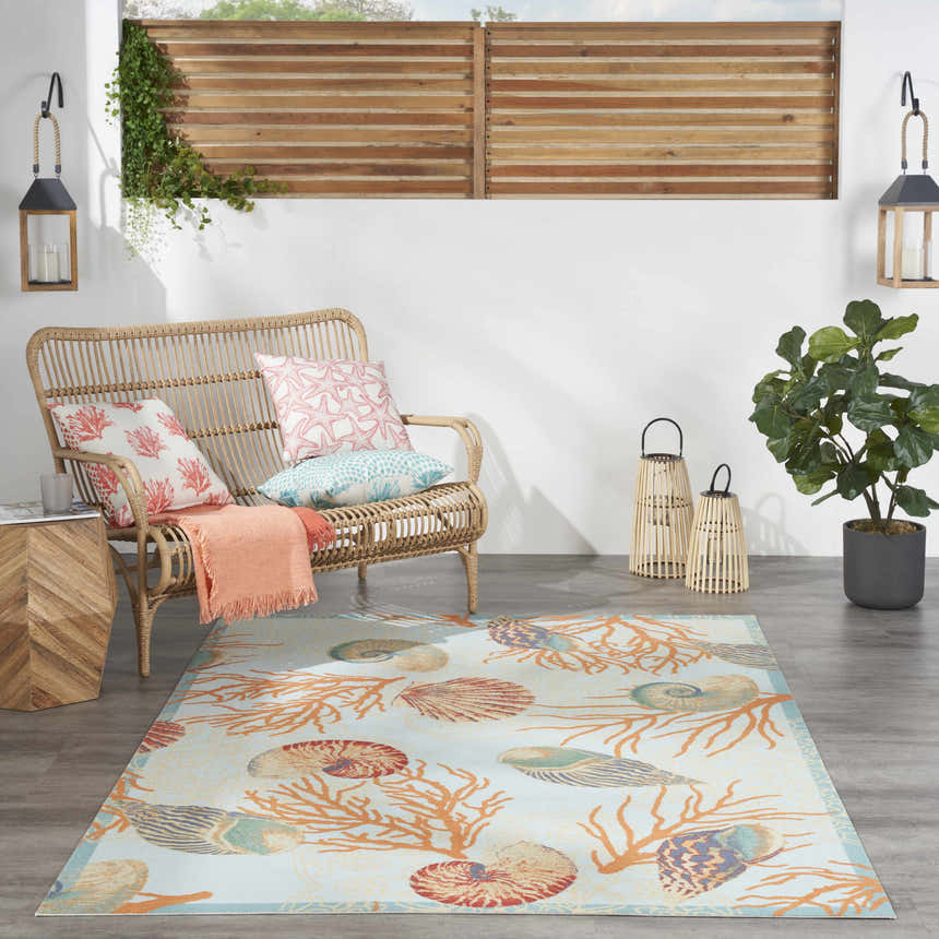 Coastal patio scene with a Waverly Sun N' Shade Light Blue rug featuring coral and seashell designs. Wicker furniture.