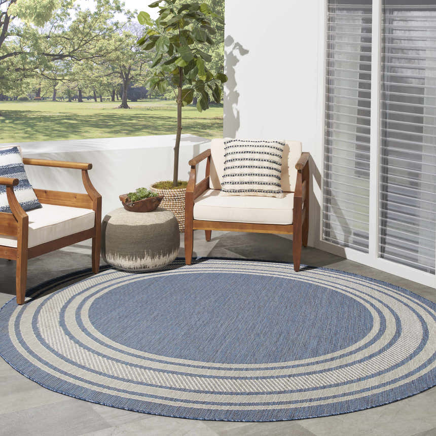 Cozy patio scene with a round, denim blue Nourison Horizon Indoor/Outdoor Area Rug, chairs, and ottoman.
