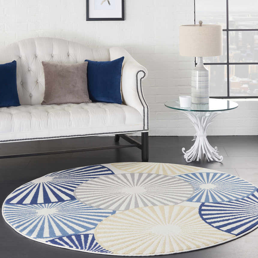 Living room scene with a round Nourison Grafix White rug, white sofa, glass table, and blue and gray pillows.