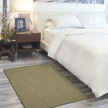 Bedroom scene with a green Nourison Courtyard rug on gray wood floors, next to a white bed and dark wood nightstand.