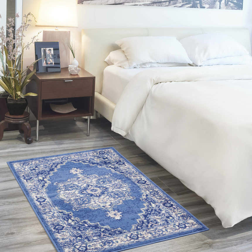Bedroom scene with a Nourison Whimsicle Navy Farmhouse Indoor Area Rug on gray wood floors. White bedding and brown nightstand.