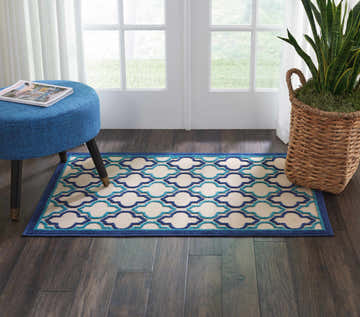 Entryway scene with a Nourison Aloha Navy Outdoor Area Rug, blue ottoman, and potted snake plant.