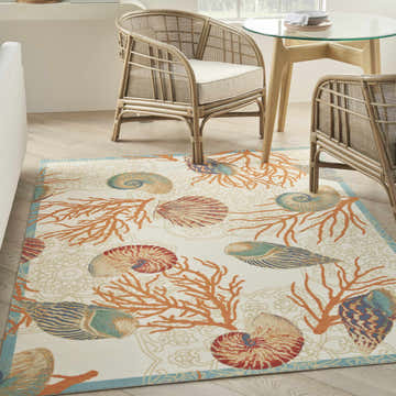Coastal dining room with Waverly Sun N' Shade rug. Ivory rug with coral and seashell pattern, rattan chairs, and glass table.