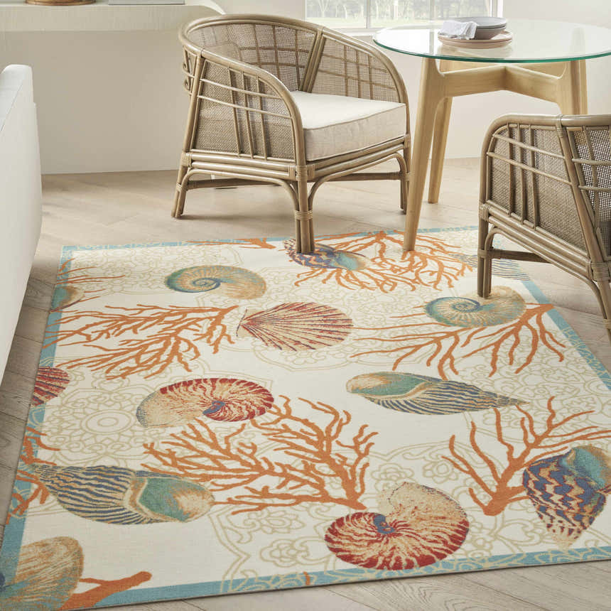 Coastal dining room with Waverly Sun N' Shade rug. Ivory rug with coral and seashell pattern, rattan chairs, and glass table.