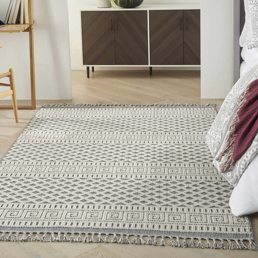 Bedroom scene with a Nourison Paxton Ivory/Slate rug, wood floors, brown cabinet, and bed with patterned throw.