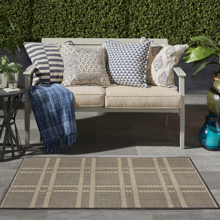Outdoor patio scene with a Nourison Horizon Charcoal rug, loveseat with blue pillows, and green foliage.