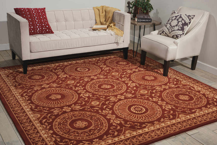 Living room scene with a brick red Nourison Versailles Palace rug, cream sofa and chair, and wood side table.