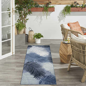 Patio scene with denim Nourison Suncoast Tropical Area Rug runner, wicker chairs, and lush plants.
