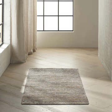 Minimalist room scene with Calvin Klein Mesa Hematite rug on light wood floor. Neutral curtains and natural light.