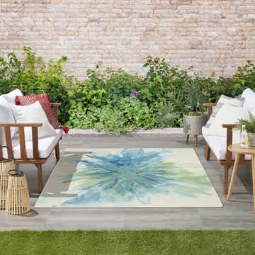 Outdoor patio scene with Nourison Color Splash Ivory Blue Modern Area Rug, wood sofas, and lush greenery.