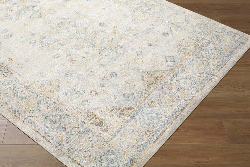 Close-up of the Bhavya Cream Washable Area Rug. Beige rug with muted blue and sage traditional pattern on a wood floor. Corner