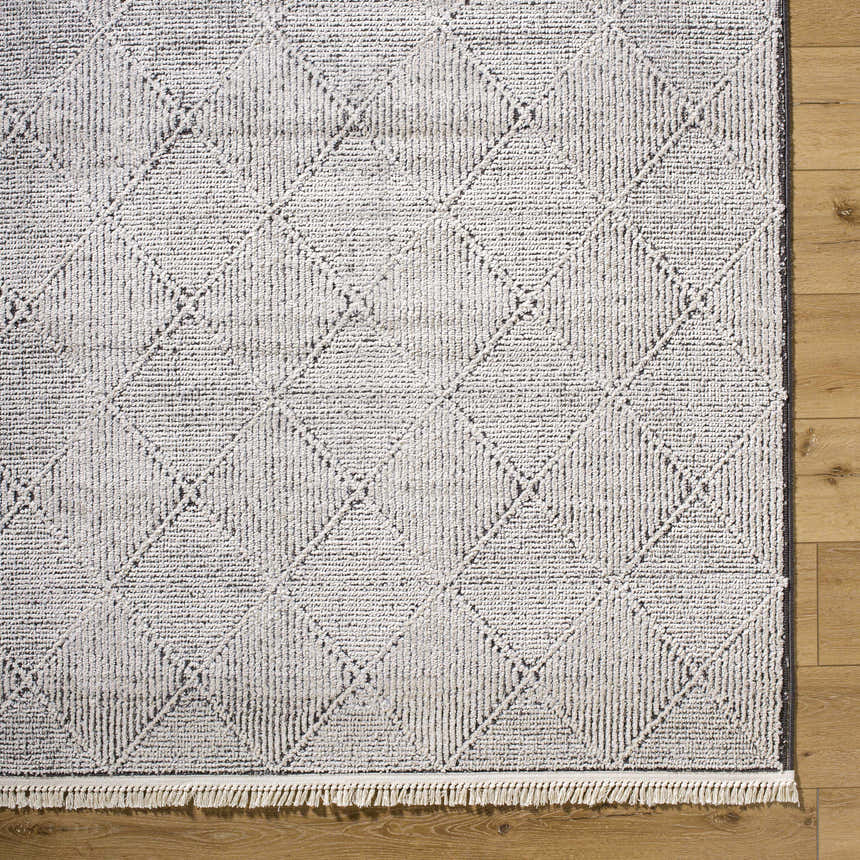 Close-up of Cannan Area Rug. Ivory rug with black geometric diamond pattern and fringe detail on wood floor. Corner