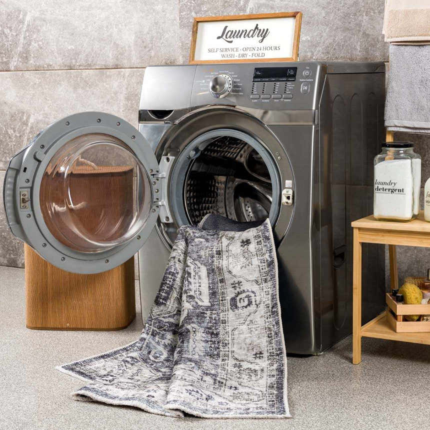 Laundry room scene with Off White Holi-4602 washable rug partially inside the washing machine. Beige, charcoal, and gray tones. Washing-Machine