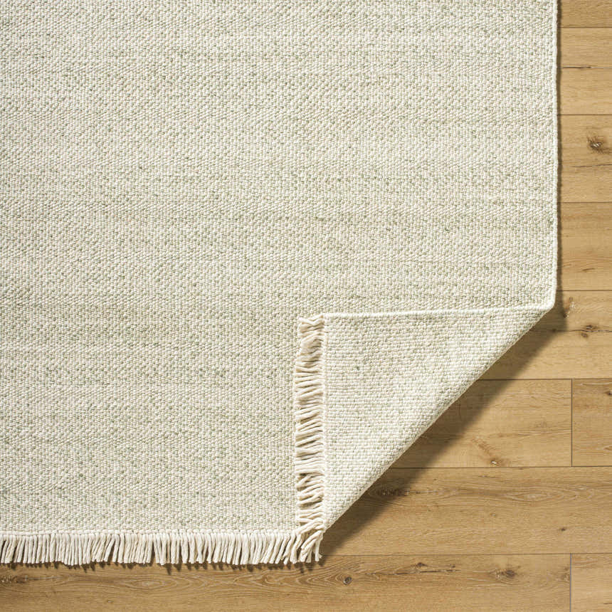 Close-up of the Sorell Sage Rug, showing its textured weave, ecru and sage colors, and wool fringe on wood floor. Fold