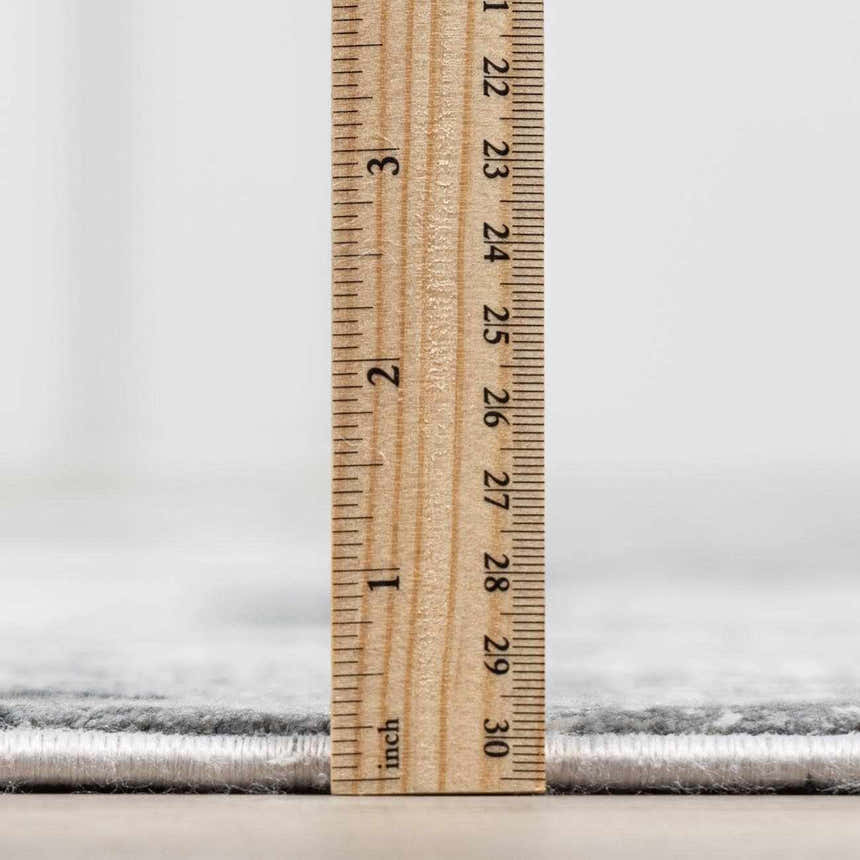 Close-up of the Littleport Luxury Fringed Faux Silk Rug showing the rug's pile height with a ruler. Gray tones. Pile