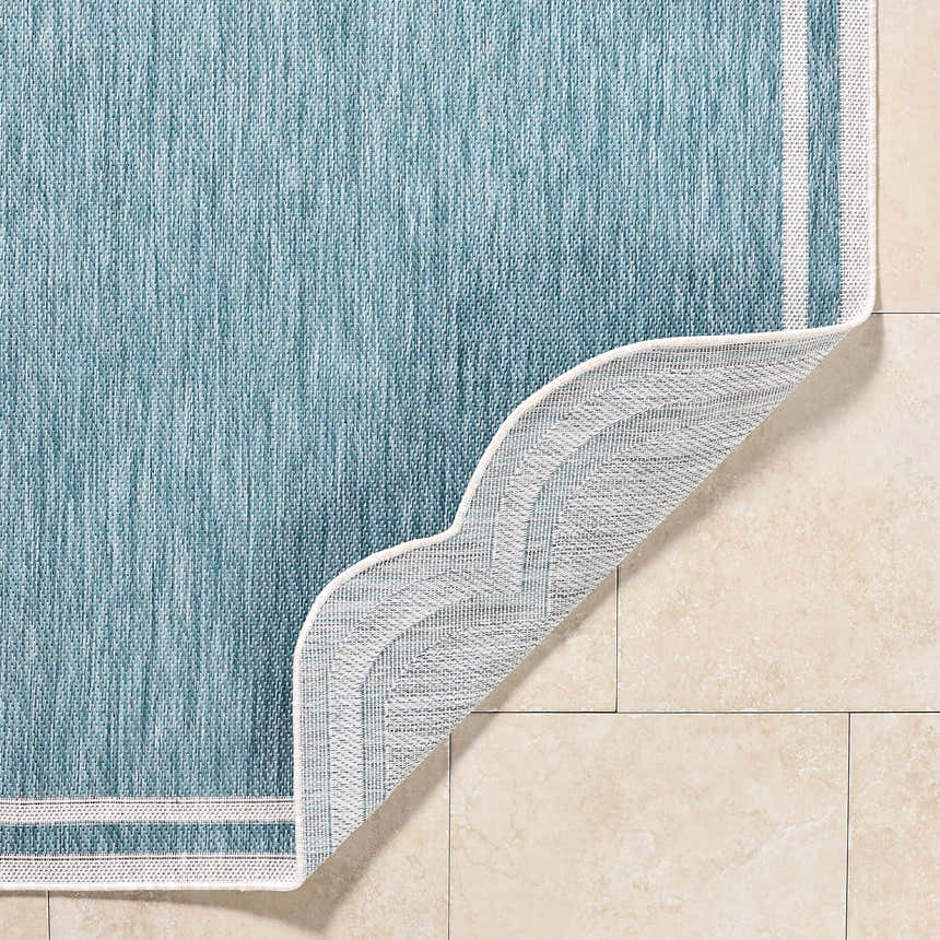 Close-up of the Arnola Light Blue Outdoor Rug showing its aqua color, woven texture, cream border, and backing. Fold