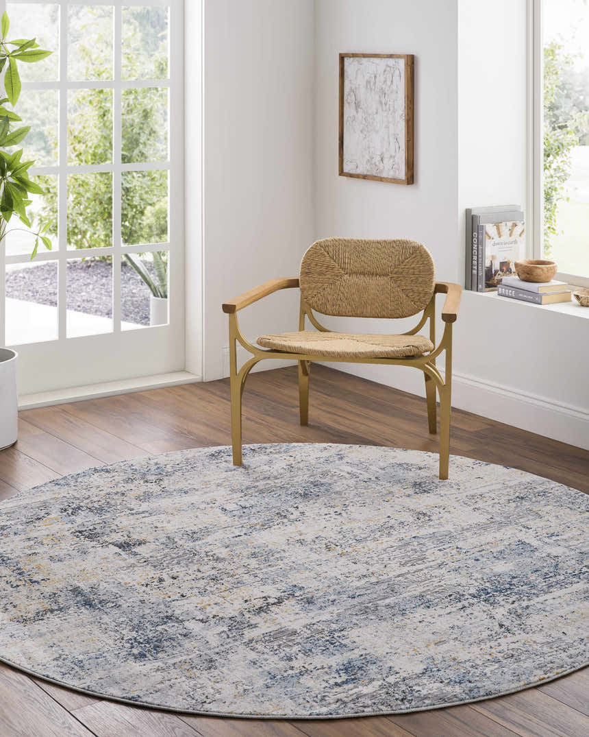 Bright room scene with a round Hyman Area Rug in gray, denim, and camel tones under a woven chair. Roomscene-Round