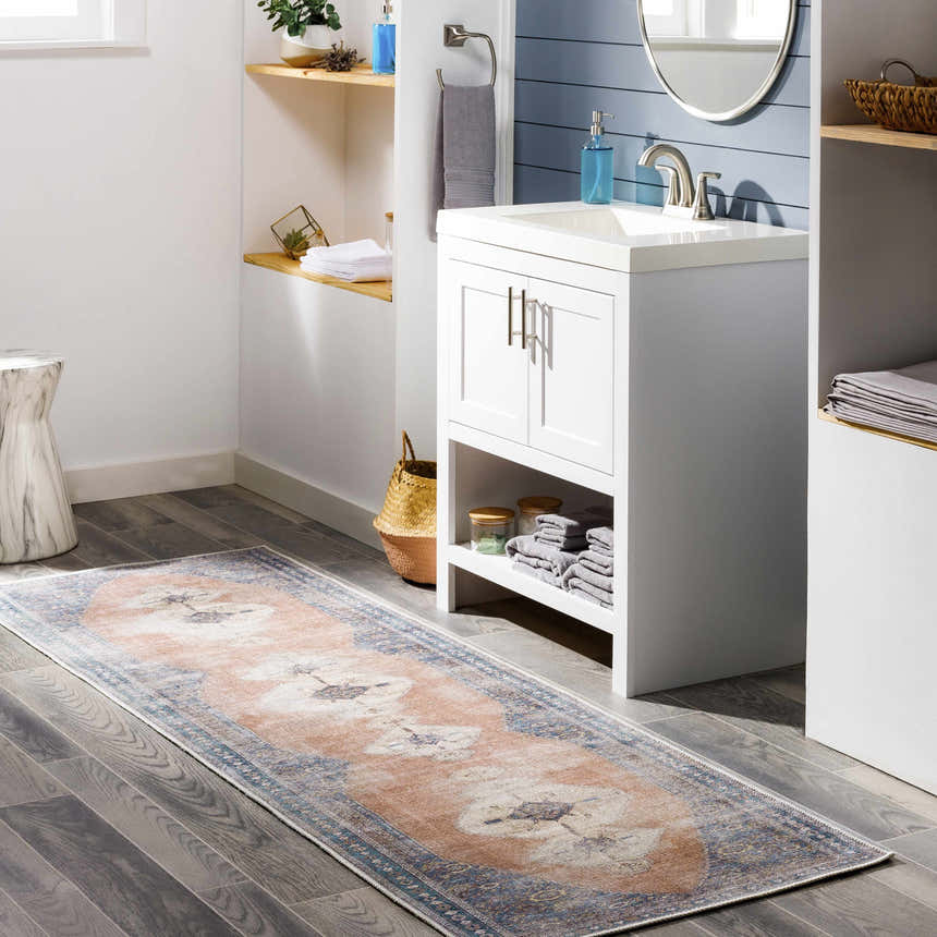Light blue bathroom scene with peach and navy Stakeford Washable Area Rug, white vanity, and gray wood floors. Roomscene-Runner