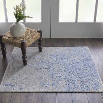 Living room scene with Nourison Silk Shadows Grey Blue rug, woven stool, vase, and dark wood floors.