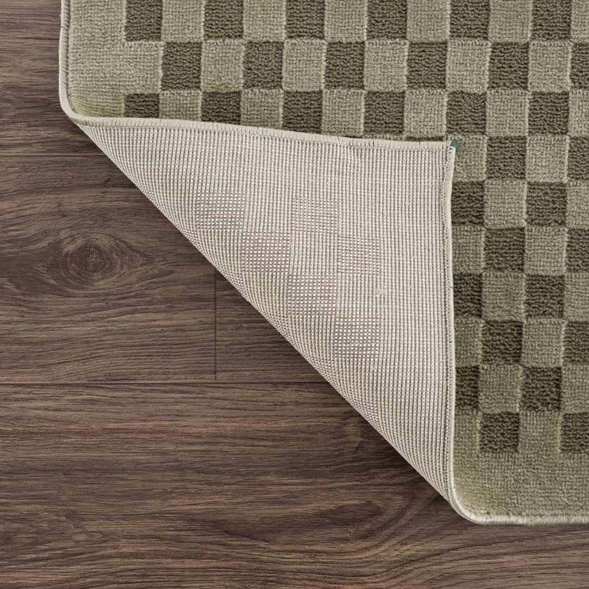 Close-up of Gaiwan Olive Green Washable Area Rug showing checkerboard pattern, texture, and backing on wood floor. Fold