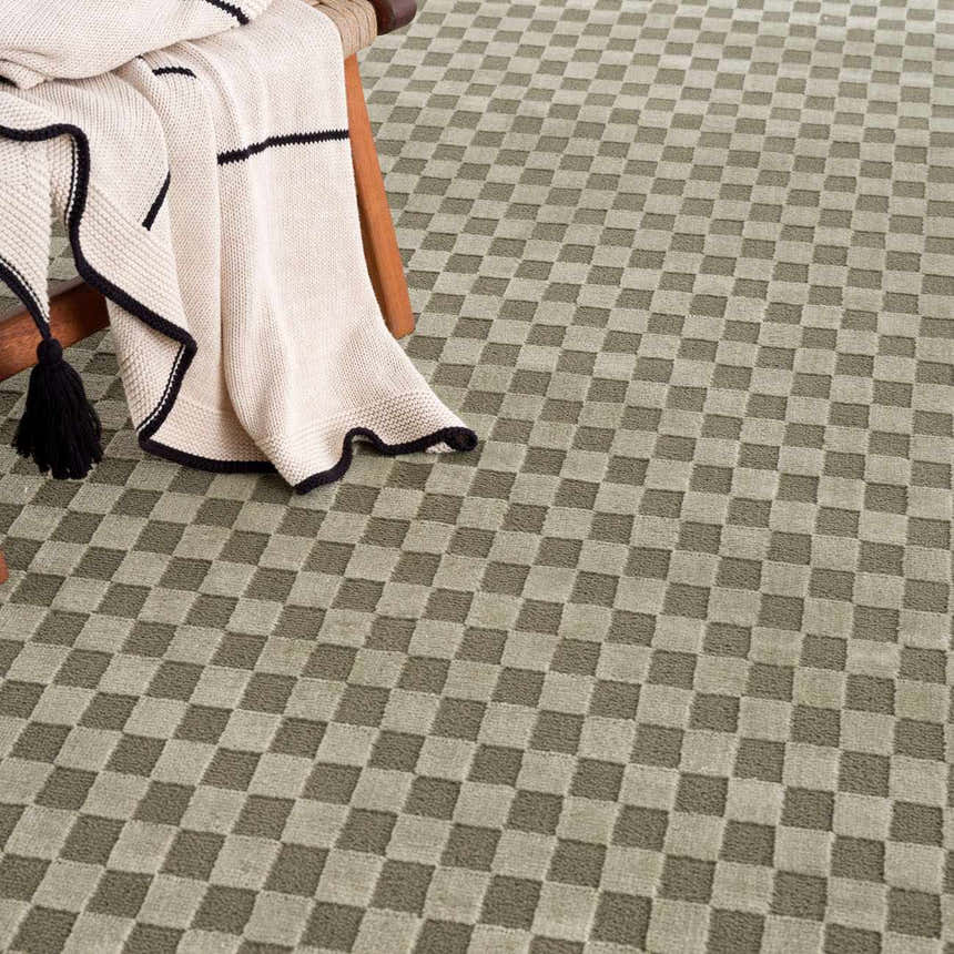 Close-up of the Gaiwan Olive Green Washable Area Rug, a gray green checkerboard pattern. Blanket on a wooden chair. Styleshot