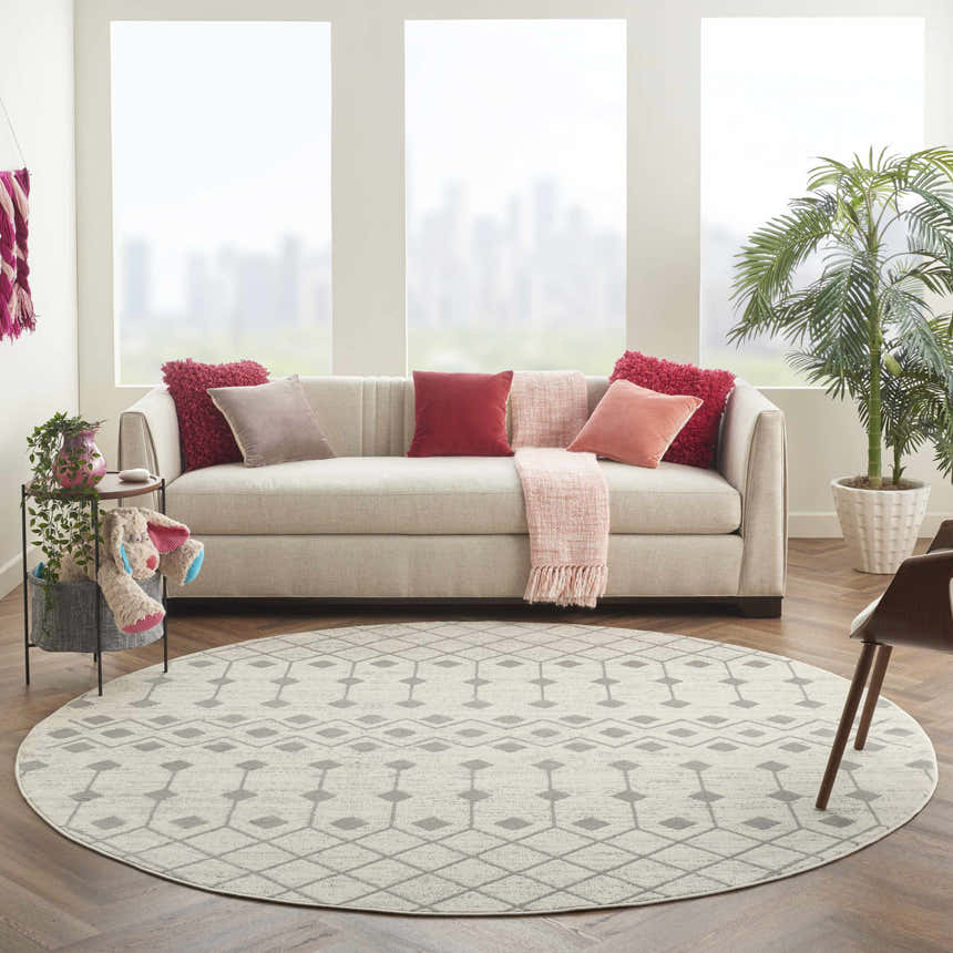 Living room scene with a round Nourison Grafix Ivory/Grey rug, neutral sofa, pink accents, and herringbone wood floors.