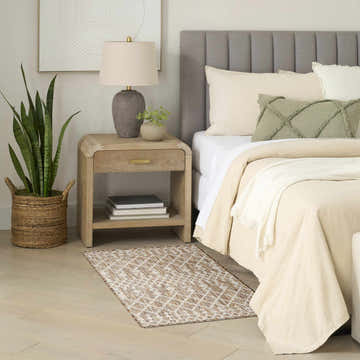 Beige bedroom scene with a Nourison Positano Beige Modern Area Rug, wood nightstand, and neutral bedding.