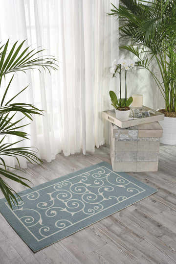 Light blue Nourison Home & Garden rug with white swirl pattern in a bright, airy room with plants and sheer curtains.