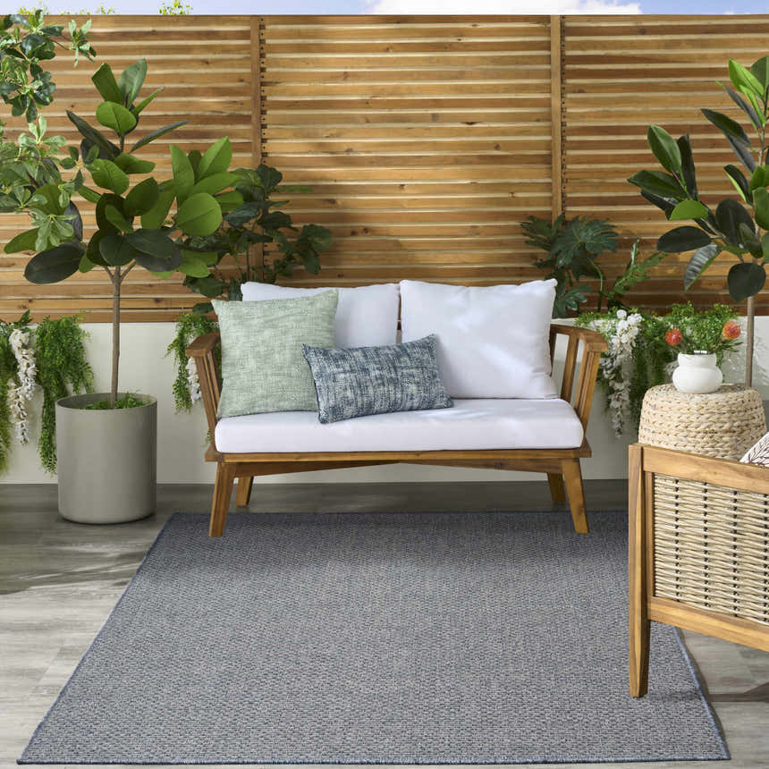 Serene patio scene with a denim Nourison Tulum Outdoor Area Rug, wood furniture, and lush greenery.
