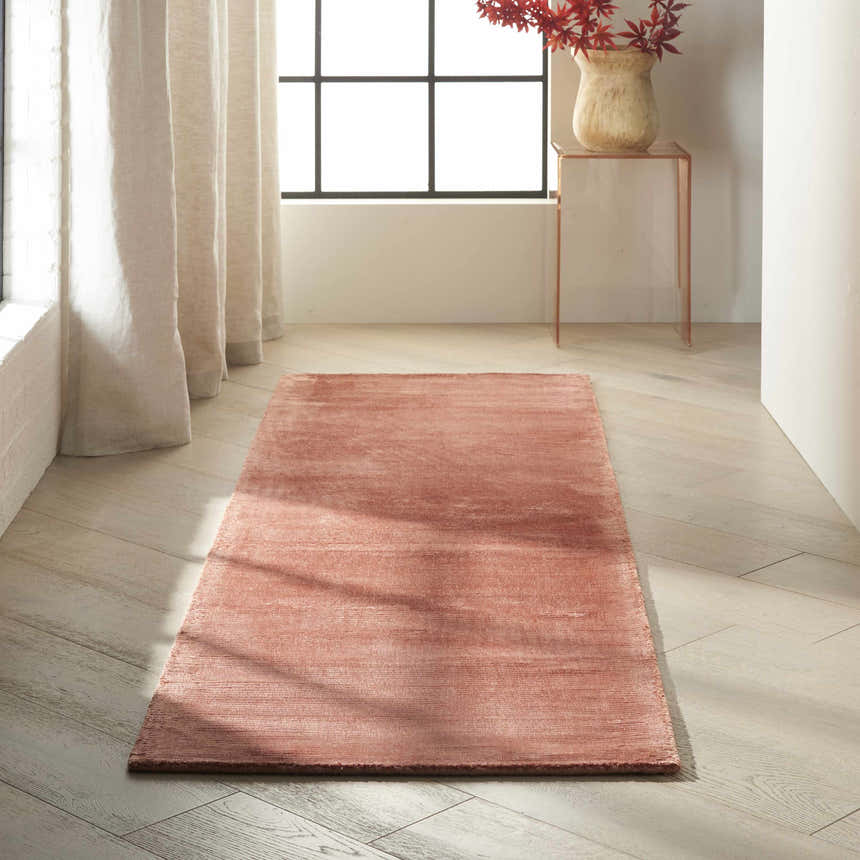 Sunlit room scene with a Calvin Klein Home Lunar Rust runner rug on light wood floors. Neutral curtains and vase with red leaves.