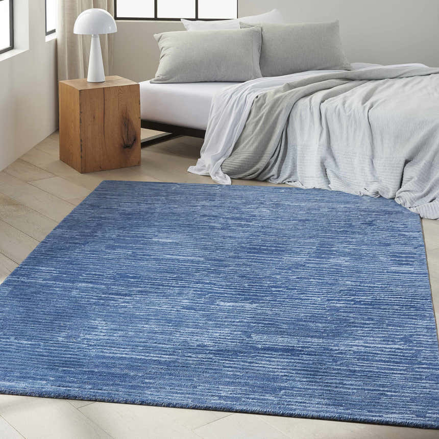 Bedroom scene with a Calvin Klein CK010 Linear Blue rug, wood floors, and a bed with gray pillows.