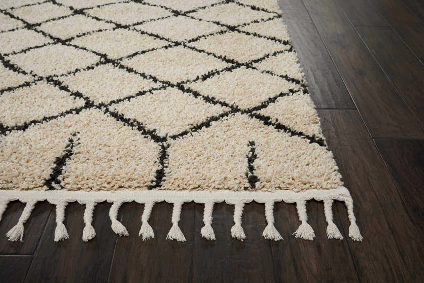 Close-up of Nourison Moroccan Shag Cream Tribal Rug with diamond pattern and fringe on dark wood floor. Soft, textured rug.