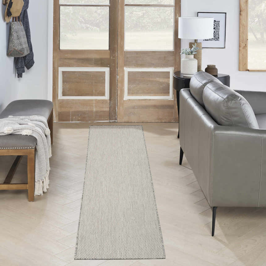 Entryway scene with a Nourison Courtyard Ivory Silver rug, gray leather couch, wood door, and herringbone wood floors.