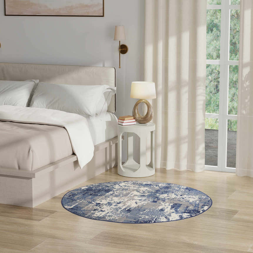 Bedroom scene with a round Nourison Grafix Navy Blue rug, bed, white nightstand, lamp, and neutral decor.