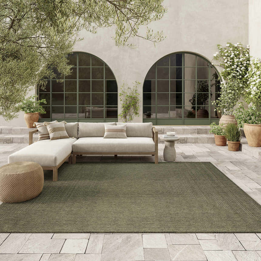 Olive Nourison Tulum outdoor rug on a stone patio with a neutral sectional sofa and potted plants.