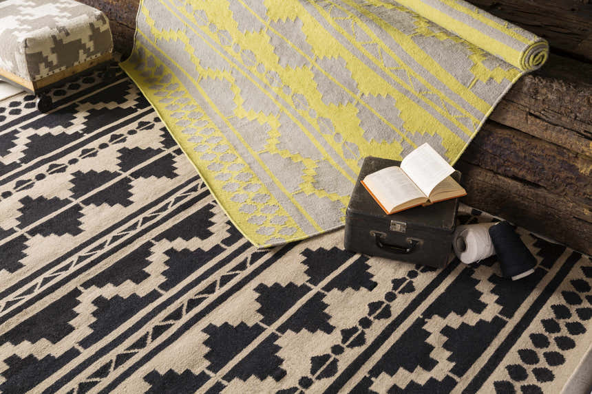 Styled Gratiot Area Rug scene: black and beige rug, rolled gray and yellow rug, trunk, yarn, and ottoman. Styleshot
