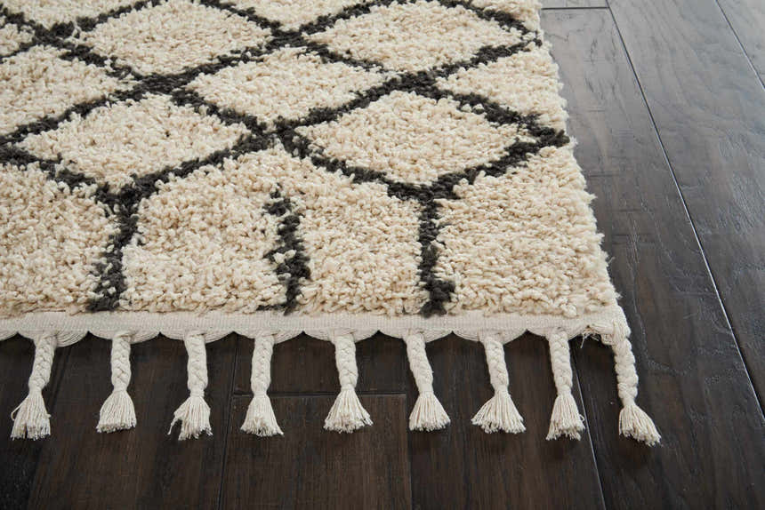 Close up of Nourison Moroccan Shag Cream Tribal rug, showing shag texture, diamond pattern, and fringe on dark wood floor.