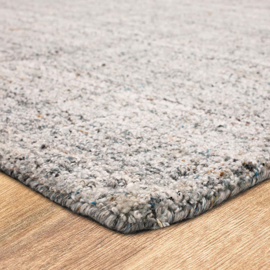 Close-up of the Karastan Haberdasher Silver Area Rug. The rug has a textured design with gray, ivory, and blue flecks. Corner