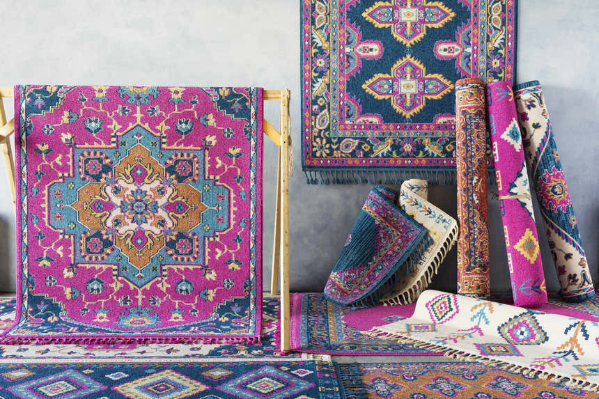Display of colorful Oban Traditional Rugs in camel, coral, blue, fuchsia, and ivory, rolled and hung against a gray wall. Styleshot