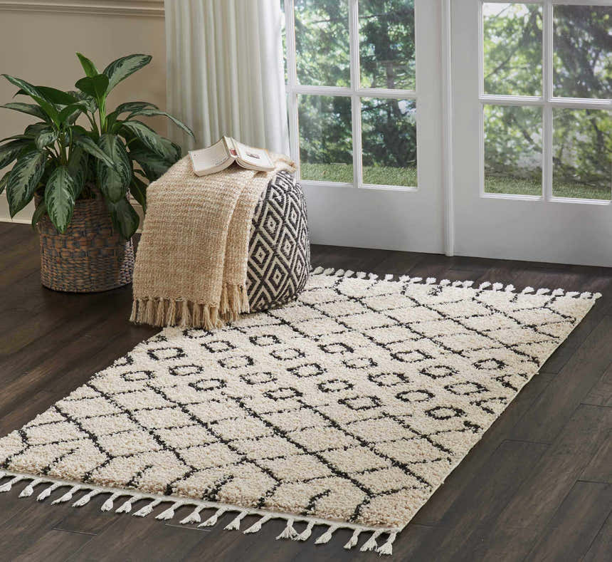 Cozy room scene with a Nourison Moroccan Shag Cream Tribal rug, pouf, blanket, and plant near a window.
