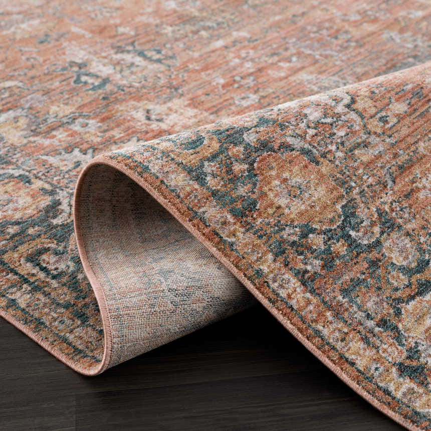 Close-up of the Mele Area Rug showing its traditional design, bone, burnt orange, and teal colors, and machine-woven texture. Fold