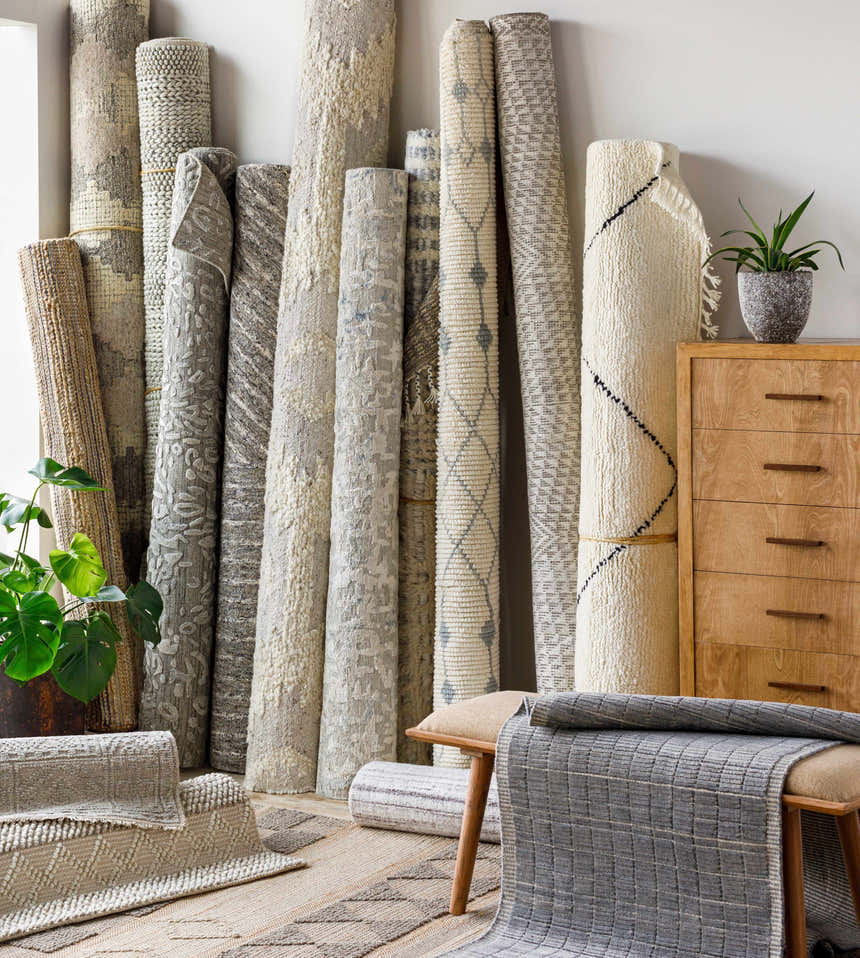 Rolled Beykoz Area Rugs in cream, beige, gray, and brown, displayed with a wood dresser, bench, and potted plant. Front