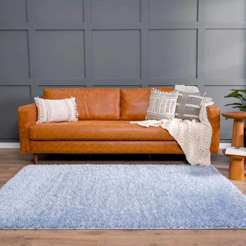 Living room scene with a Faina Solid Aqua Shag Rug, tan leather sofa, throw pillows, wood floors, and gray paneled wall. Roomscene-Rectangle