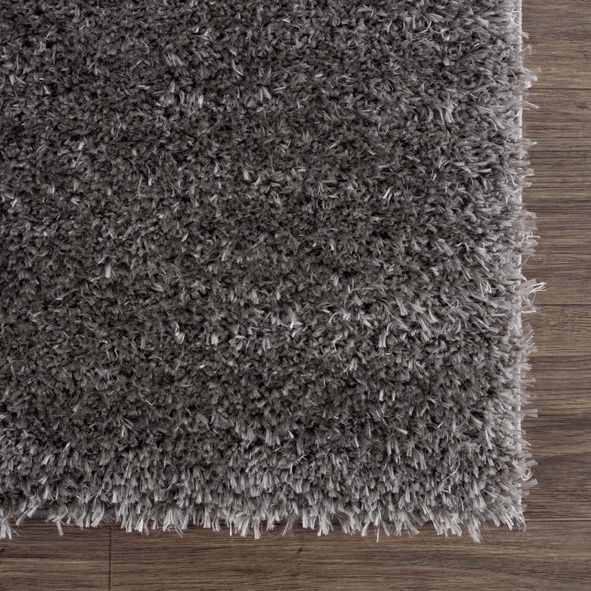 Close-up of the plush texture of the Faina Solid Dark Gray Shag Washable Area Rug on dark wood flooring. Swatch